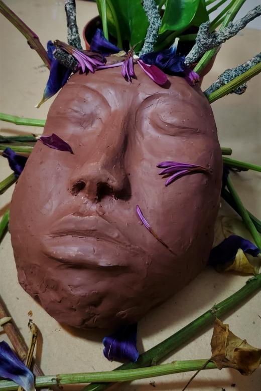 Close up of a terracotta colored clay sculpture of a face with green potho leaves sprouting from the top of the ahead along with sticks, flower petals, cut flower stems, and dried leaves.