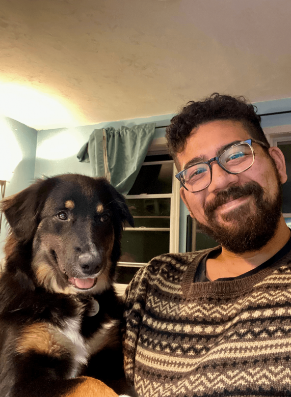 Selfie of Andrew and a black and brown dog. Andrew smiles while wearing a sweater and glasses.
