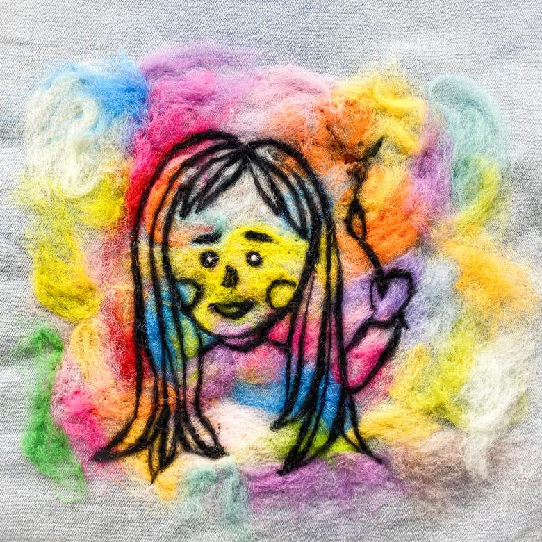 A white square piece of cloth with a rainbow blurred felt square shape in the center with a black threaded outline of a girl smiling with long hair holding up a paint brush