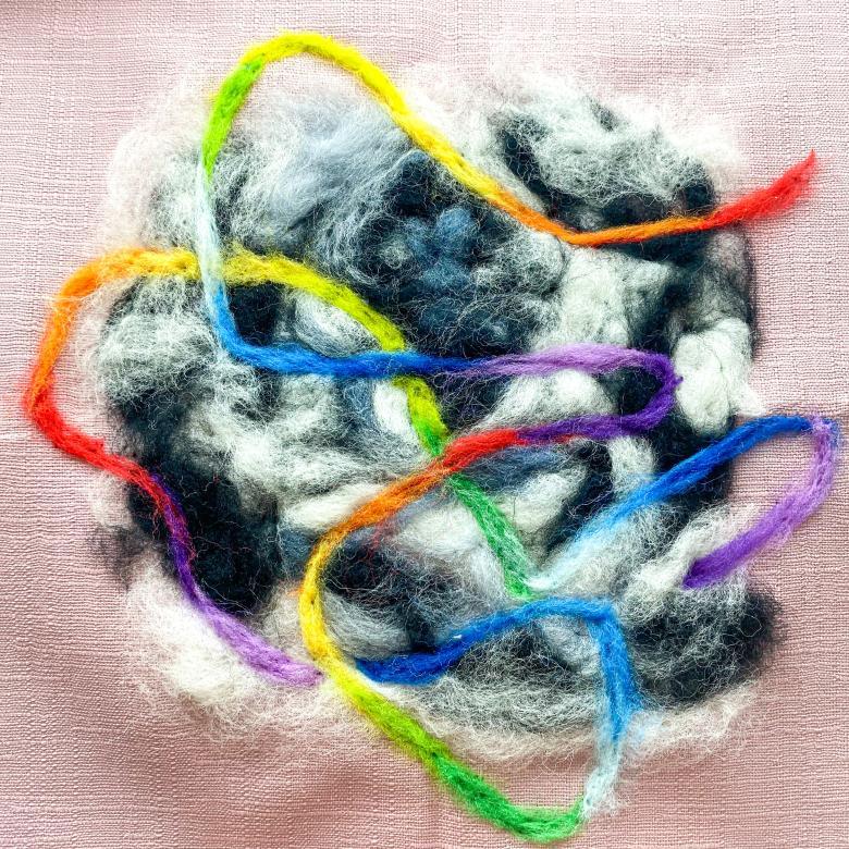 A light pink fabric square with a black, white, and grey swirled felt circular mass in center. Through the circle is a thin rainbow line that swirls in and out of the mass. 