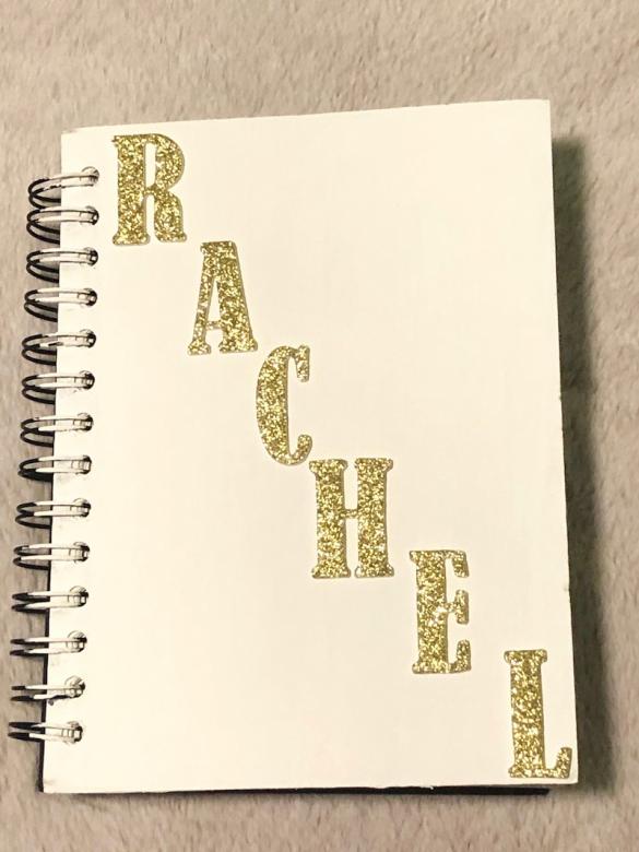 The cover of the small spiral bound notebook, painted white, with gold lettering that reads “RACHEL”