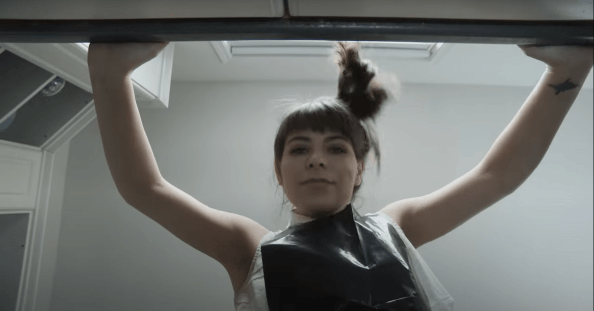 Woman with bangs and a ponytail looks down at the camera while leaning on a kitchen counter. She is smiling slightly.