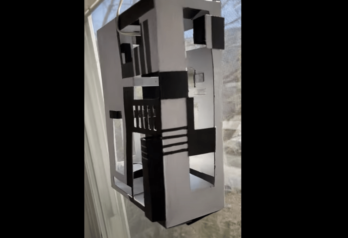 Video still of a white and grey small cardboard building with lots of windows and doors cutout mounted onto a real window overlooking trees and a blue sky.