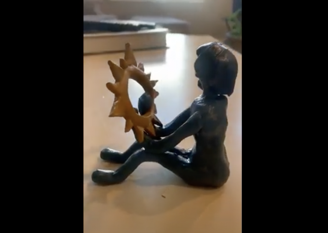 Video still of a dark blue sculpture of a figure sitting holding a bright yellow moon/sun like shape on the knees with rays coming out.  