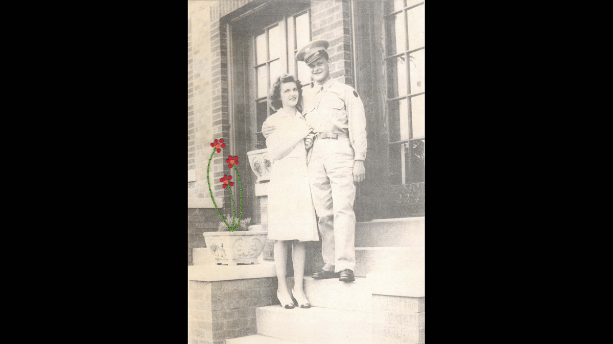 Black and white vintage photograph of a man in uniform with his arm around a woman. They stand on steps outside a building. Three red and orange flowers with green stems are sewn on top of the photograph growing out of a pot beside the couple.