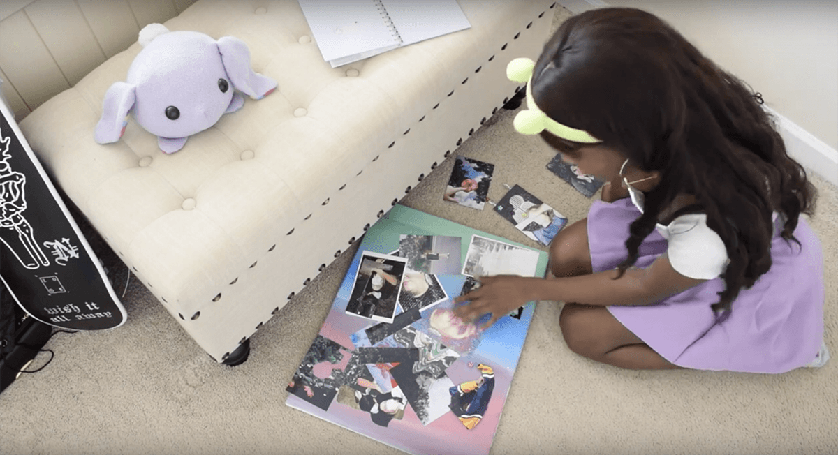 Looking down at a young Black girl with long brown hair, a green headband, a white shirt, and a purple skirt. She is bent down working on a photo collage.