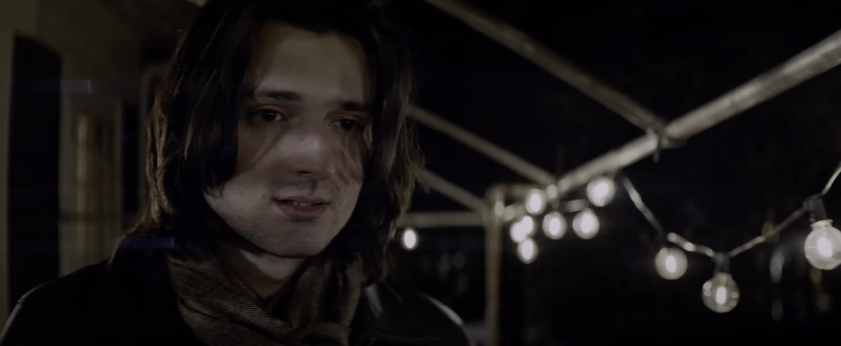 Close-up of a man with shoulder-length hair looking off-camera. It is night and he stands on a porch with a string of bulbs hanging from the roof's overhang. 