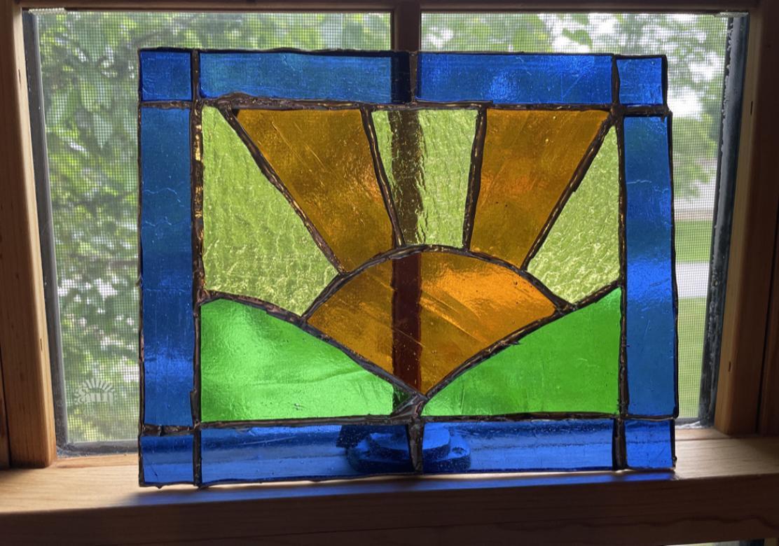 Photograph of a stained glass rectangle featuring an orange sun positioned between two green hills. Orange and yellow sunbeams radiate out of the sun. Royal blue stained glass creates a rectangular border. This photo shows the piece in a sunny window. 