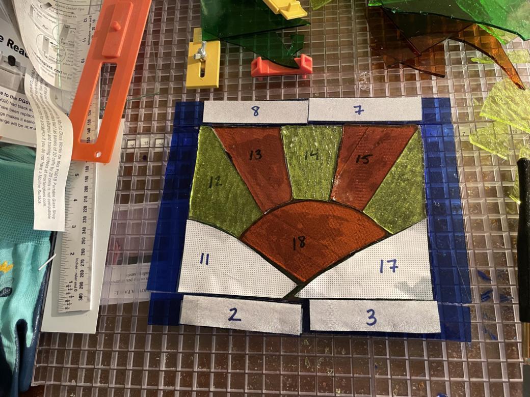 aturing an orange sun positioned between two green hills. Orange and yellow sunbeams radiate out of the sun. Royal blue stained glass creates a rectangular border. In this progress photo, the glass is on a cutting mat with various tools. White numbered cut outs of paper are positioned on the glass so the artist can cut and piece the glass.