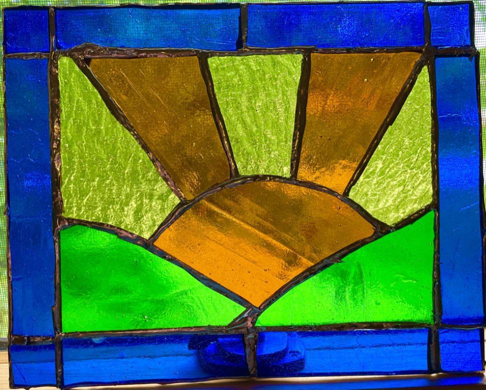 Photograph of a stained glass rectangle featuring an orange sun positioned between two green hills. Orange and yellow sunbeams radiate out of the sun. Royal blue stained glass creates a rectangular border. 
