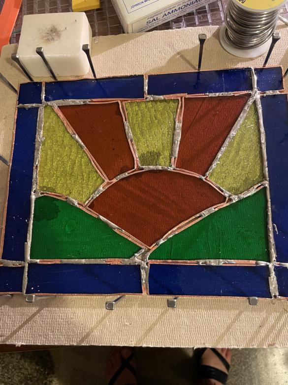 Photo of a stained glass sunset in orange and yellow peaking through green hills with a royal blue rectangular border. In this image the artwork is placed on a worktop space with silver nails around the edges while the metal melts. 