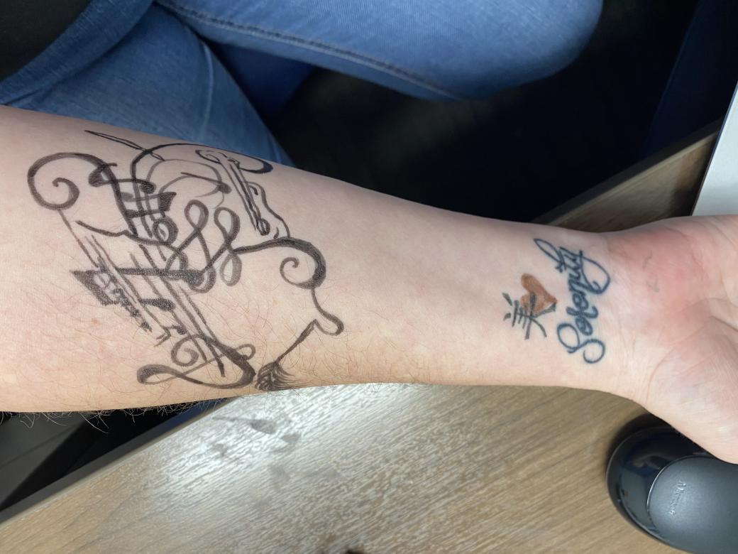 Vertical photograph of a human forearm facing up showing a black ink drawing of musical notes, swirls, and symbols. This is a drawing of the artist's image "Come Together 2". Lower down on the arm towards the wrist is a tattoo of a red heart and character above the word serenity in script font.