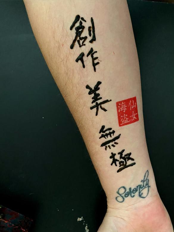 Black henna ink detailing Chinese calligraphy characters in a vertical line with a red square character to the right center. This is featured on a human forearm turned facing up on a black background.