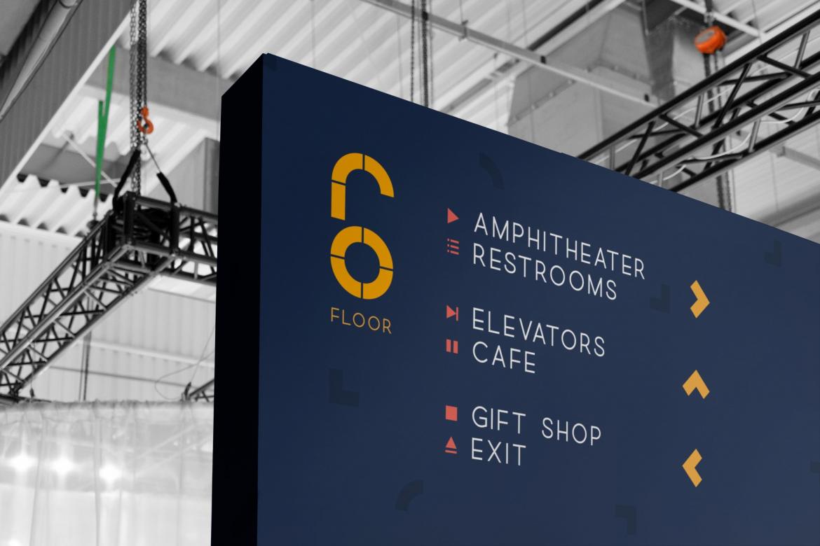 A large sign hanging from the ceiling that matches Madalena's geometric branding. The sign says "6 Floor" and gives directions to the amphitheater, restrooms, elevators, cafe, gift shop, and exit.