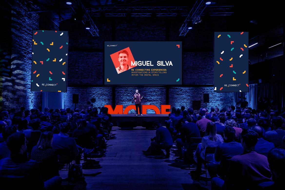 Dark room with blue lighting. Rows of people in chairs face a speaker on stage. Graphics of the MODE branding decorate the screens behind the speaker.