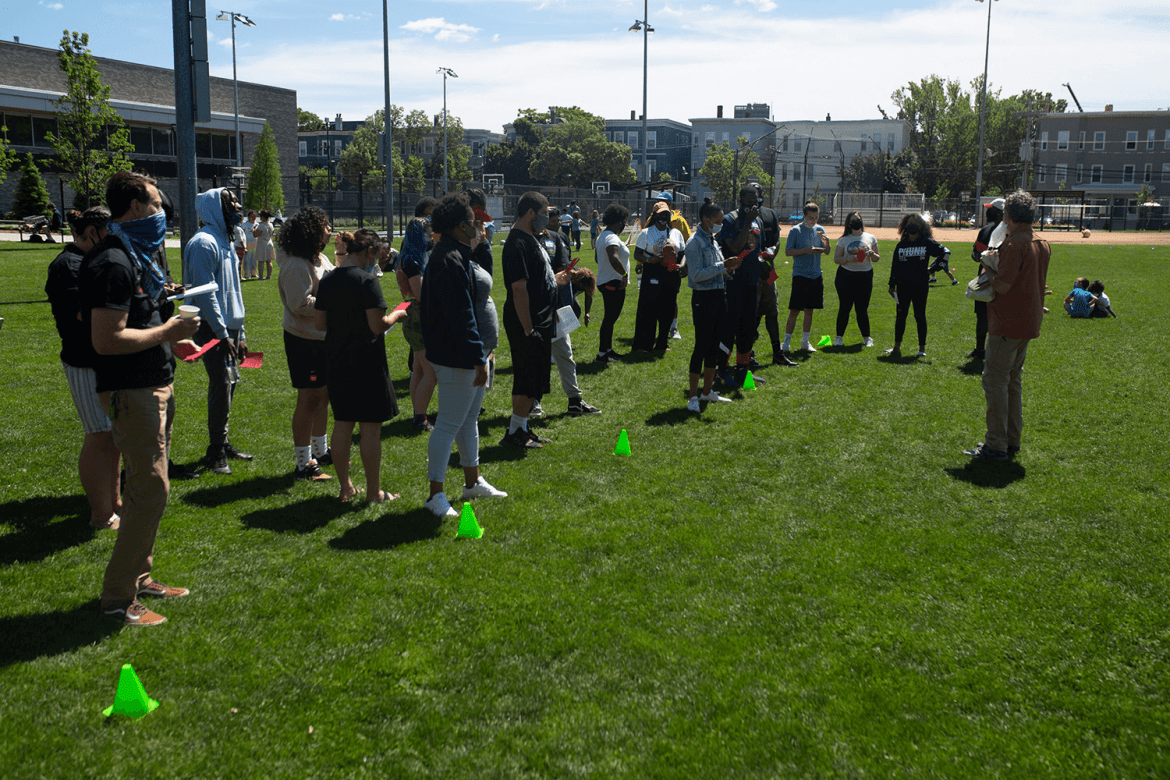 A large group of people standing on a baseball field, forming lines behind small green cones.