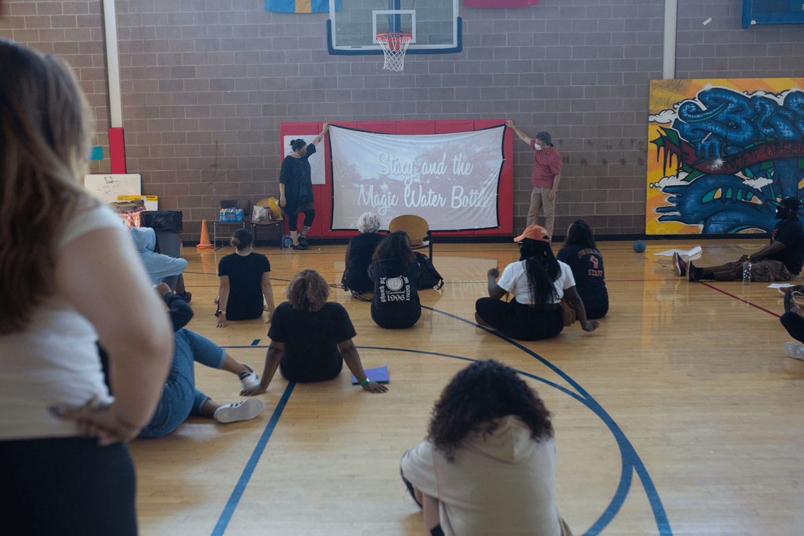 A group sitting socially distant in a school gym watch a screen that two people are holding up. A projection on the screen shows text that says "Stacy and the Magic Water Bottle."
