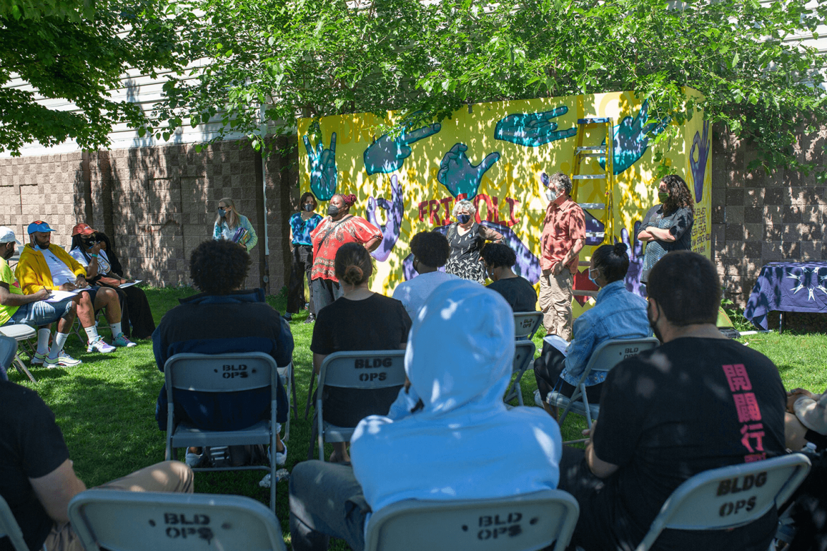 People sit outside in metal folding chairs facing a line of people who stand in front of a painted mural. The mural says "FRISOLI" and the word is surrounded by painted hands. 