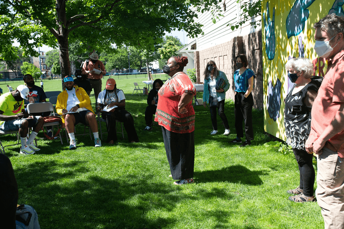 Outside a woman with her hands on her hips stands in front of a group of people sitting in metal folding chairs filling out paperwork.