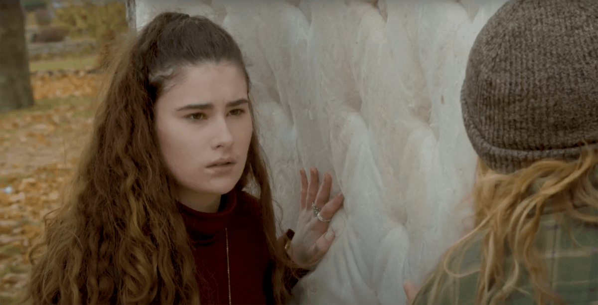 A girl with long curly hair looks angrily at another girl who's back is to us. The girl with curly hair has her hand against a mattress.