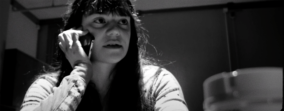 Black and white image of a girl with long hair and bangs. She sits at a table and holds a cellphone to her ear.