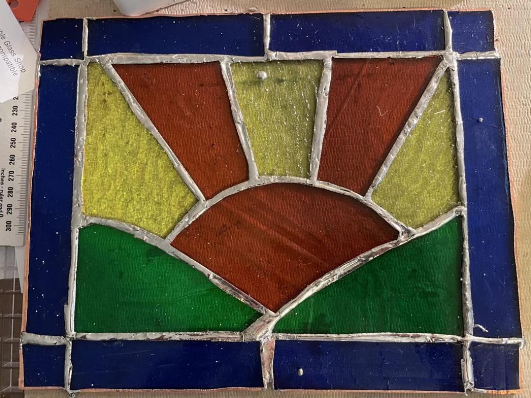 Photograph of a stained glass rectangle featuring an orange sun positioned between two green hills. Orange and yellow sunbeams radiate out of the sun. Royal blue stained glass creates a rectangular border. 
