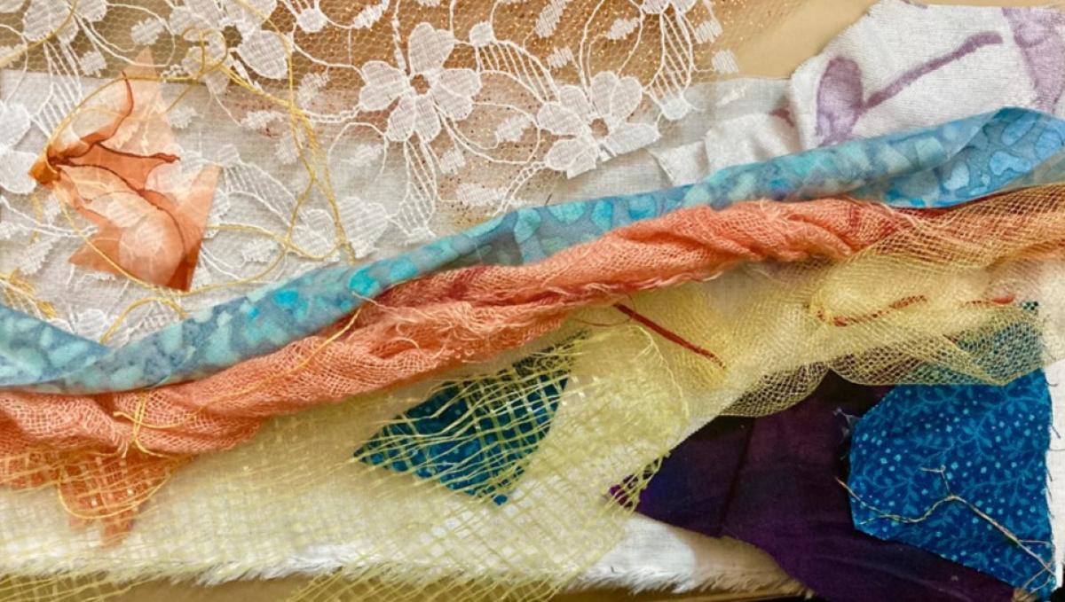 Mixed Media. Several pieces of fabric overlay each other on a yellow background. Assorted colors of fabric are twisted and create a horizontal composition. The colors include blue, yellow orange, brown, and white. Underneath three quarters of the fabric, starting at the bottom is a white piece of fabric with a frayed edge. The fabrics vary in texture and print including lace and mesh.