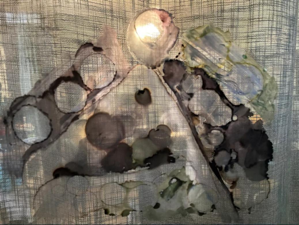 The background of this piece is a curtain with the sunshining through. The shape of a mountain is dark purple and green. The surface of the mountain is filled with purple/green circles. On the edges of the mountain are large strips of purple with circles punched out of it. On the top right is a strip of green with two large circles punched out of it. At the peak of the mountain is another purple circle with a hole punched out of the center. This hole is lined up with the sun which is shining through.