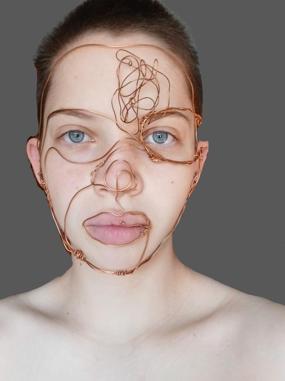 Photograph and Wire Sculpture. A person from the shoulders up with short light brown hair and light blue eyes. They are looking straight on into the camera with a straight face. Their face is wrapped up in copper wire which outlines their features.