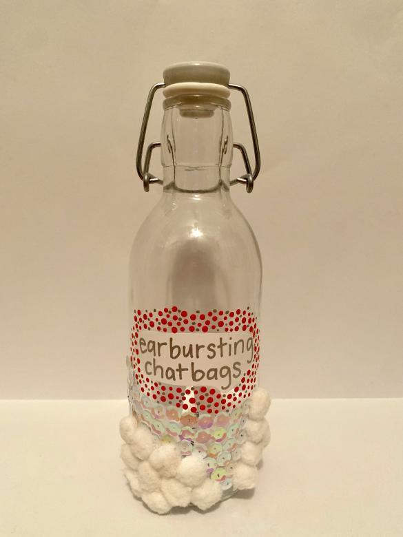 Mixed media. A glass bottle with a built-in stopper. On the bottle is a piece of paper fixed to it with the words “earbusting chatbags” written on it in silver. Around this piece of paper are small red and grey dots. Below the words are glued white and pink sequins and white pompoms.