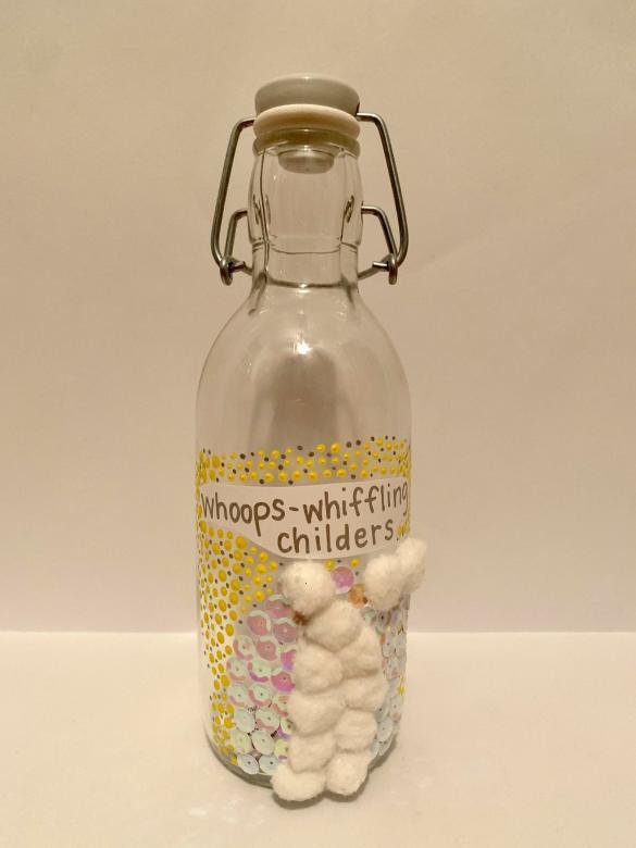 Mixed media. A glass bottle with a built-in stopper. On the bottle is a piece of paper with the words “gloriumptious human beans” written in silver. Surrounding these words are pink and white sequins and white pompoms and on the outside of those are navy blue and silver. In the bottle can be seen small slips of paper that appear to have words written on them.