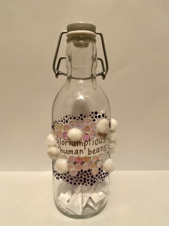 Mixed media. A glass bottle with a built-in stoppers. The bottle has a piece of paper with the words “whoops-whiffling childers” written in silver. Surrounding the words are yellow and silver dots which also extend down to surround white pompoms and pink and white sequins which are below the words.