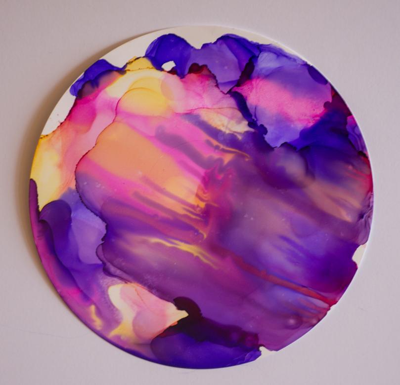 On a circular white piece of paper, are the colors orange, purple, and pink. They are mixed in many places and in some spots run together to create a drip like effect. Around the edged can be seen some of the white paper. 
