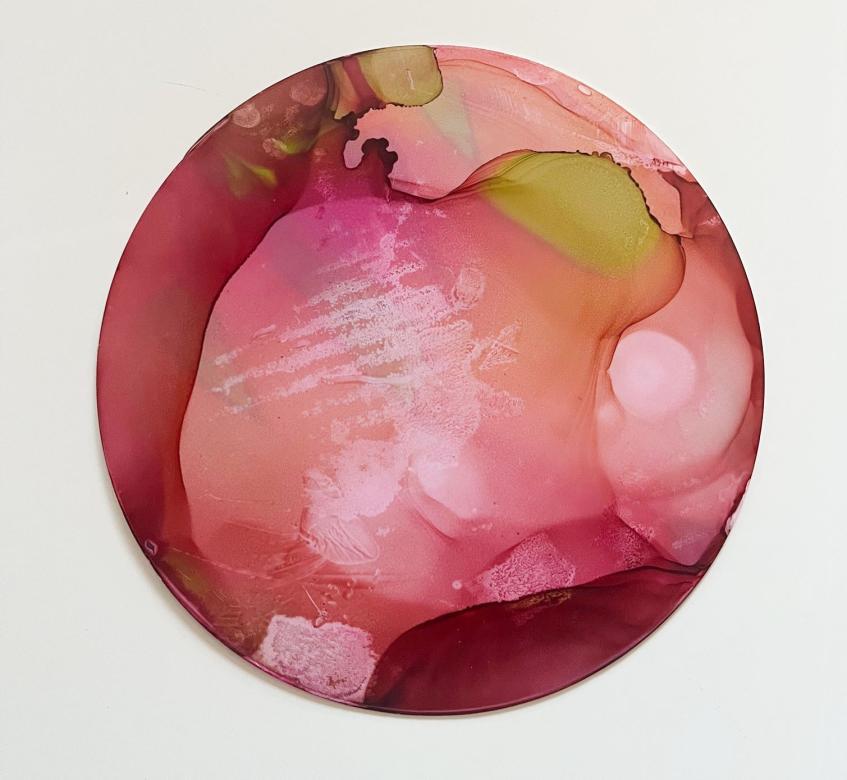  On a circular piece of paper are painted the colors maroon and red and pink. Around the edges is the maroon color and closer to the center is the pink color. At the top of the piece there are slight green accents. Over the pink and maroon colors in some spots is white drawn on in lines and splotches. 