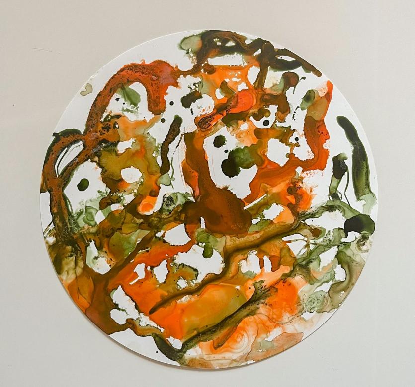 On a circular white piece of paper are the colors green and orange. They are painted in splotches of color that mix in some spots and are distinct in others. There are several gaps between these splotches where the white paper can be seen.