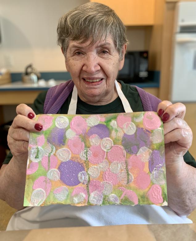 Photograph of artist Maria Carrasco smiling facing the camera holding a small colorful abstract painting.