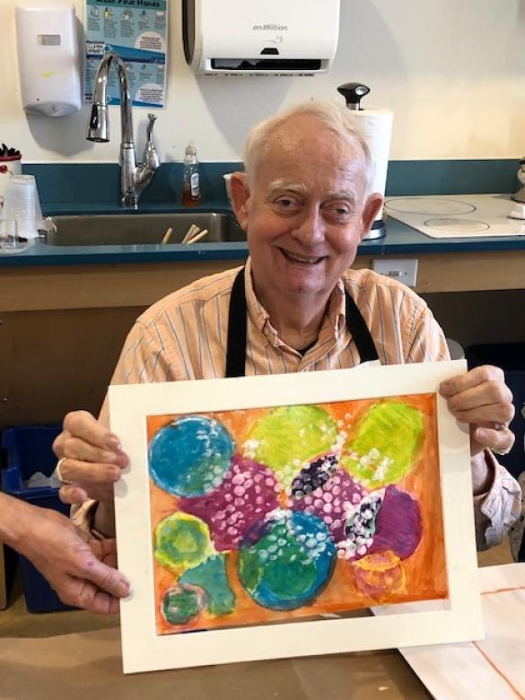 Photograph of artist Ed Jacobson smiling facing the camera holding a small colorful abstract painting.