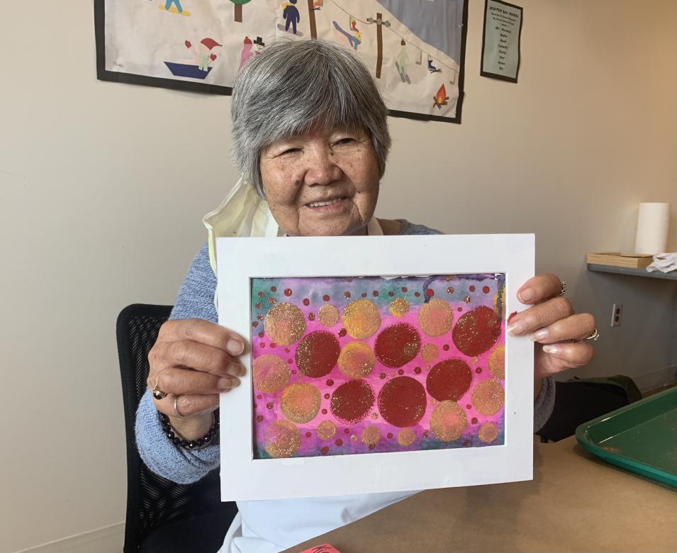 Photograph of artist Kinuko Pearce smiling facing the camera holding a small red and pink painting.