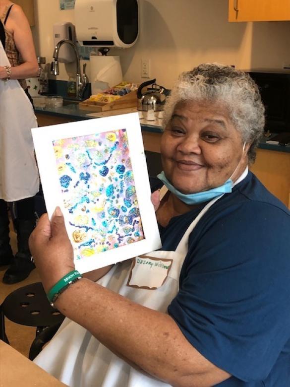 Photograph of artist Delory Williams smiling facing the camera holding a small colorful abstract painting.
