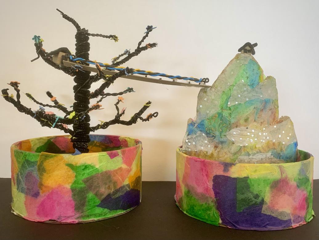 Two rainbow tissue paper circles side by side encapsulate a black pipe cleaner tree with small flowers on the left with a bridge like object to a sparkly colorful mountain on the right with a small brown house at the top.. 