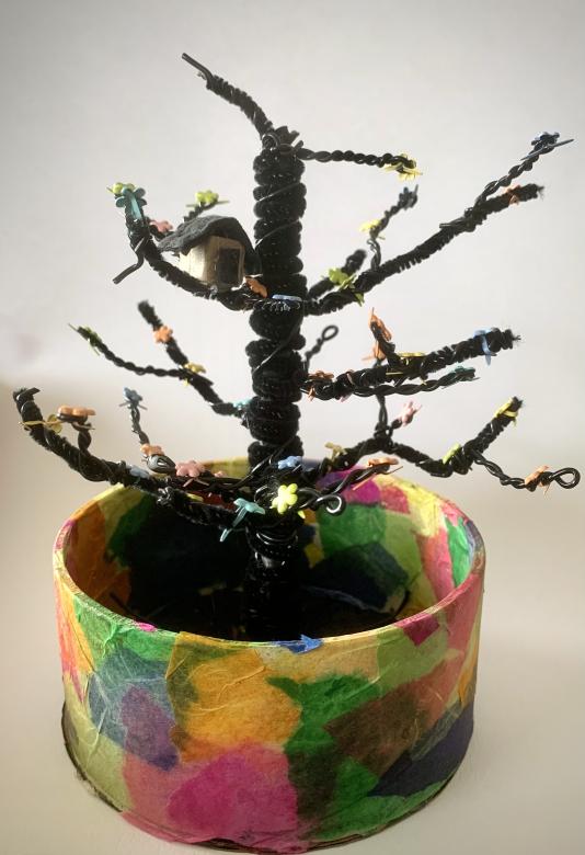 A rainbow tissue paper circle encapsulating a black pipe cleaner tree with small flowers and a tiny brown house sitting on a top branch.