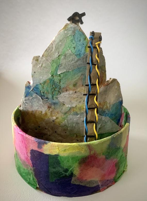 A colorful tissue paper circle encapsulating a mountain made of colorful sparkly tissue paper with textured mountain ridges. A small brown house sits on top with a wire ladder leading up.