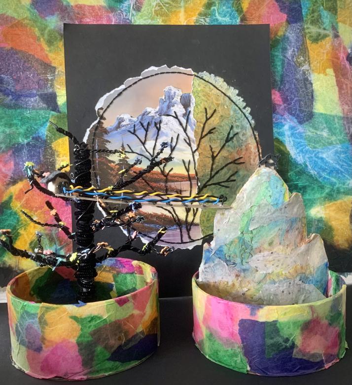 View of the tree and mountain sculptures from "Coming Home", they are now placed on a black table with a black sheet of paper in the background with a circular collage image of mountains with a black tree embroidered over. In the background is a collage of colored tissue paper.
