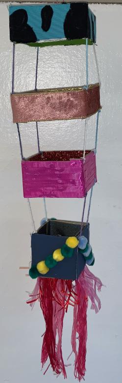 Hanging mobile featuring four distinct horizontal strips of colorful ribbon/fabric shaped into squares connected with thin string. Pom-poms adorn the final square with a variety of ribbon cascading down. 