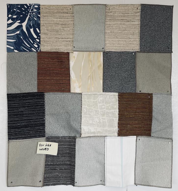 A mixed media textile piece displaying rectangular pieces of neutral gray, beige, brown, and blue fabric forming a rectangle in four rows and five columns. Each piece of fabric is stitched together with black thread. In the bottom left corner is a small note that reads "You are loved"