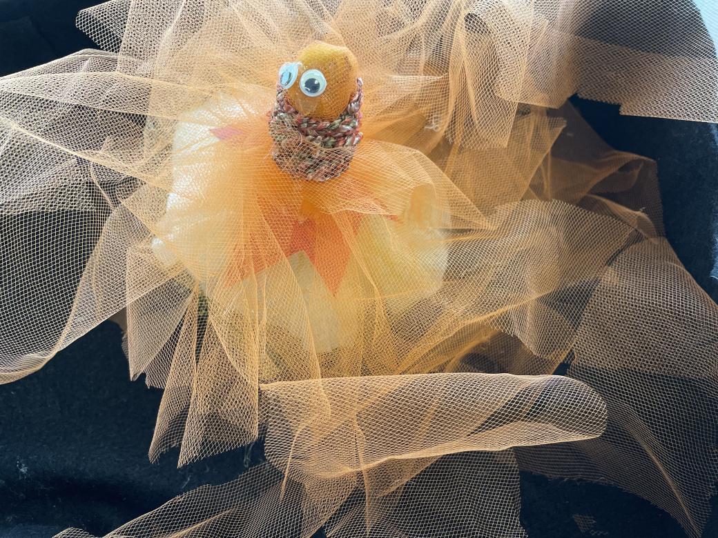 A small creature like object made from a coffee filter wrapped in orange paper and translucent orange tulle with multicolored warm toned yarn wrapped creating a neck like area, above which sits two googly eyes. 