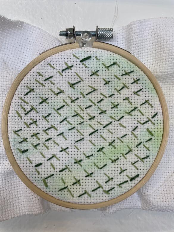 White linen cloth is stretched on a circular embroidery hoop. The linen has splotches of different shades of green watercolor. Light and dark green stitch lines repeat in diagonal lines overlapping in some areas. 