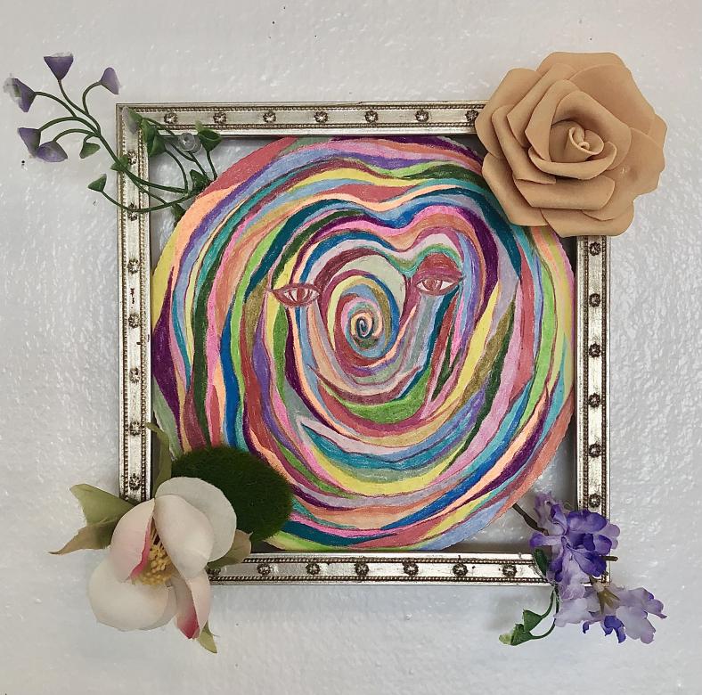 A rainbow circular drawing of swirled colors with eyes, a nose, and lips in the center. This piece is centered in a silver square frame with fake flowers in each corner. 