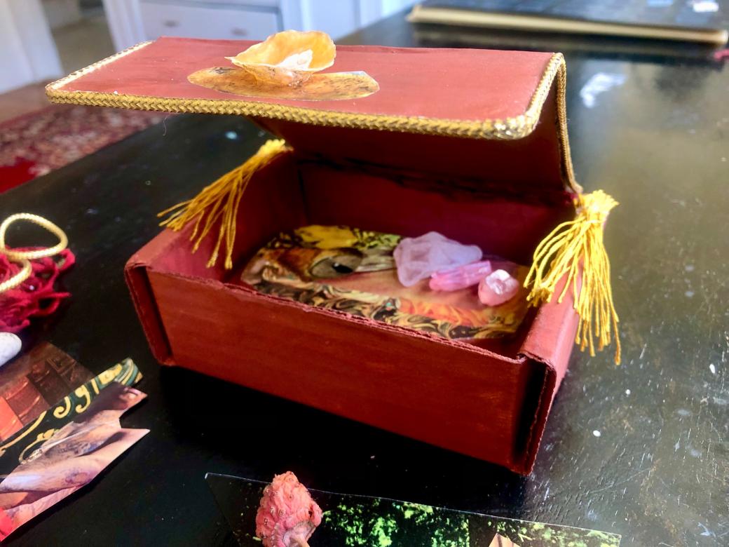 Image of a small rectangular red box with a gold rope border and yellow tassels. Inside the box is collaged images and crystals.