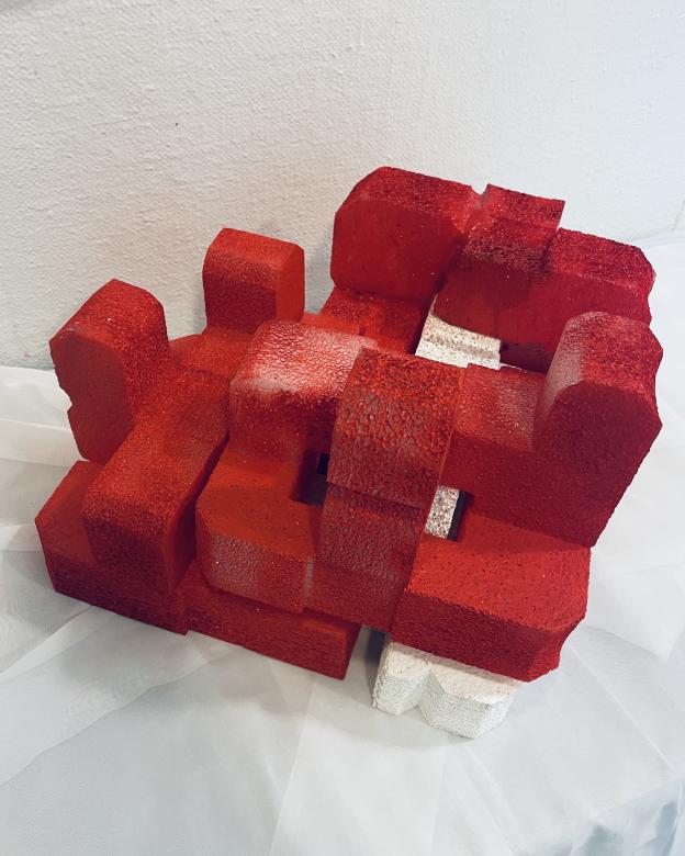 Side view of "Puzzled" a red sculpture appearing from a foam like material. The sculpture is forming a heart like shape. It has different rectangular levels appearing in different block like shapes and heights; from the side you can see through some of the shapes. In the center is a small white area.